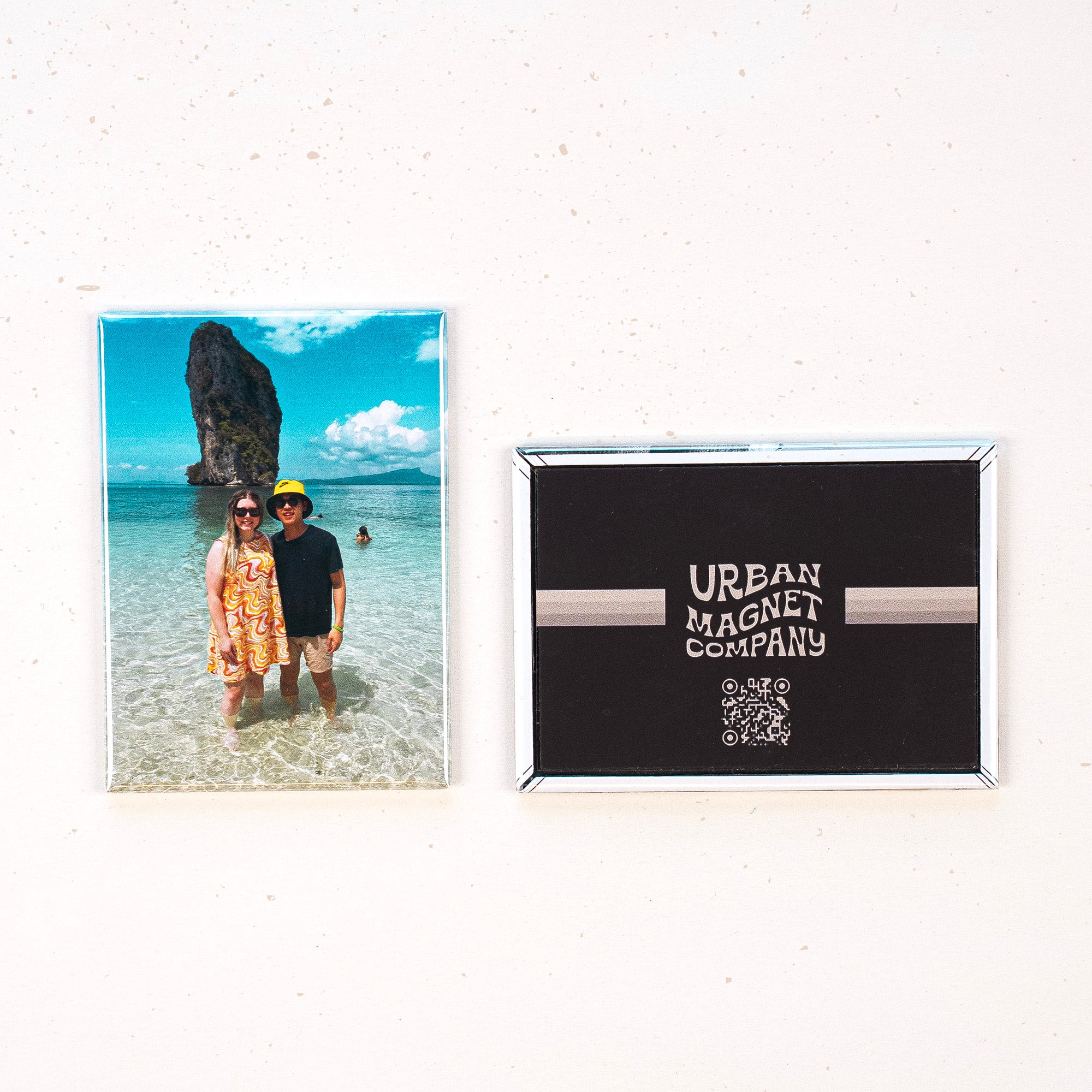 Two magnets displayed on a white background, one is printed with a photo of a couple at the beach, one is printed with the Urban Magnet Co logo. 